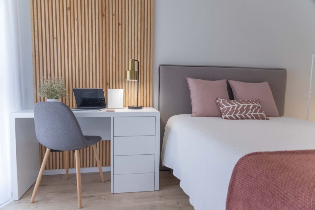 A wide horizontal interior shot of a white minimalist computer desk with drawers next to a bed and a wooden fluted slat wall panel in an aesthetic bedroom workspace.
